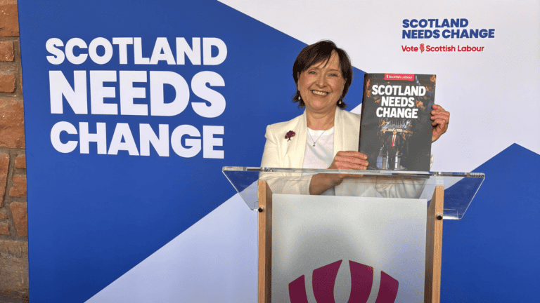 Woman holding the Scottish Labour manifesto in front of a Saltire banner saying Scotland Needs Change