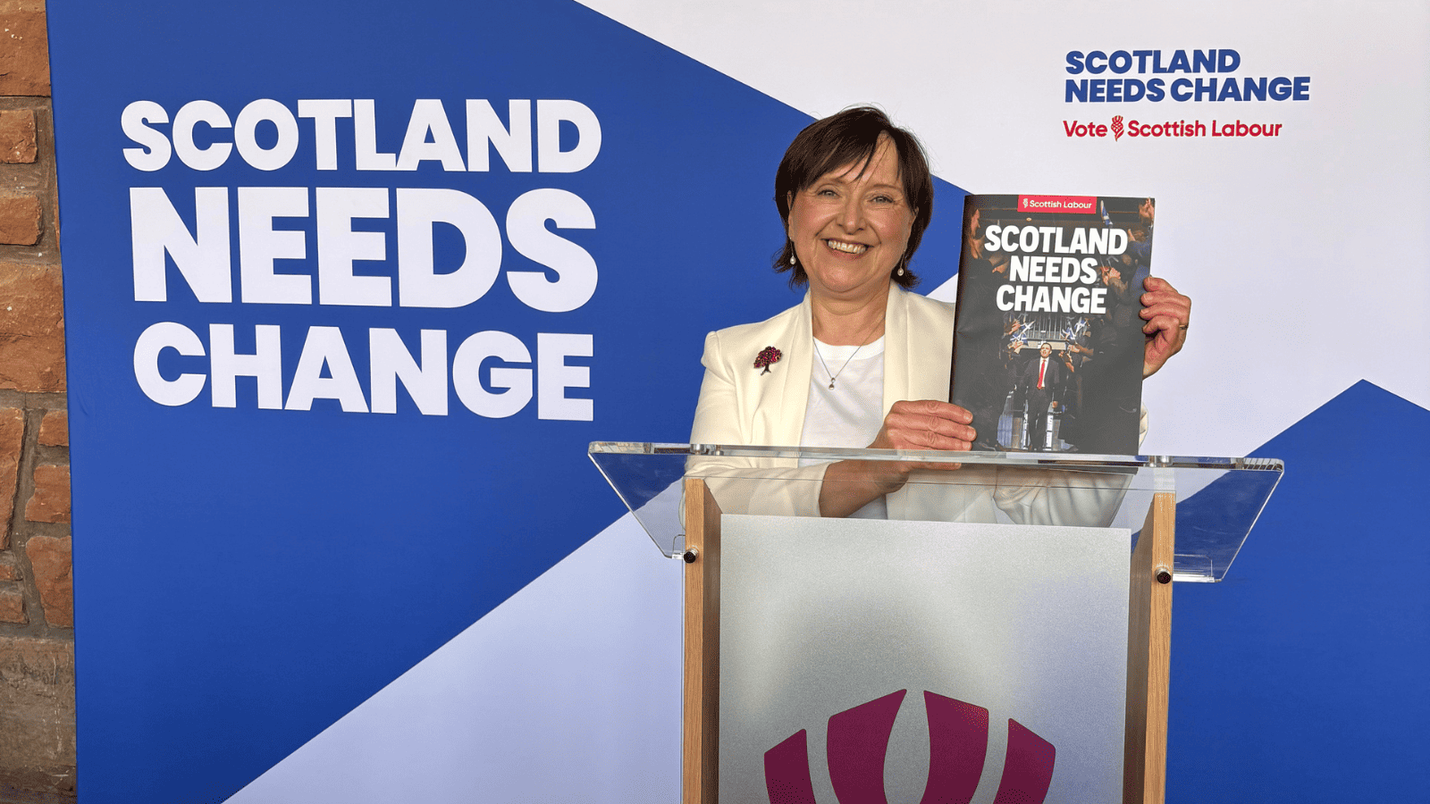 Woman holding the Scottish Labour manifesto in front of a Saltire banner saying Scotland Needs Change